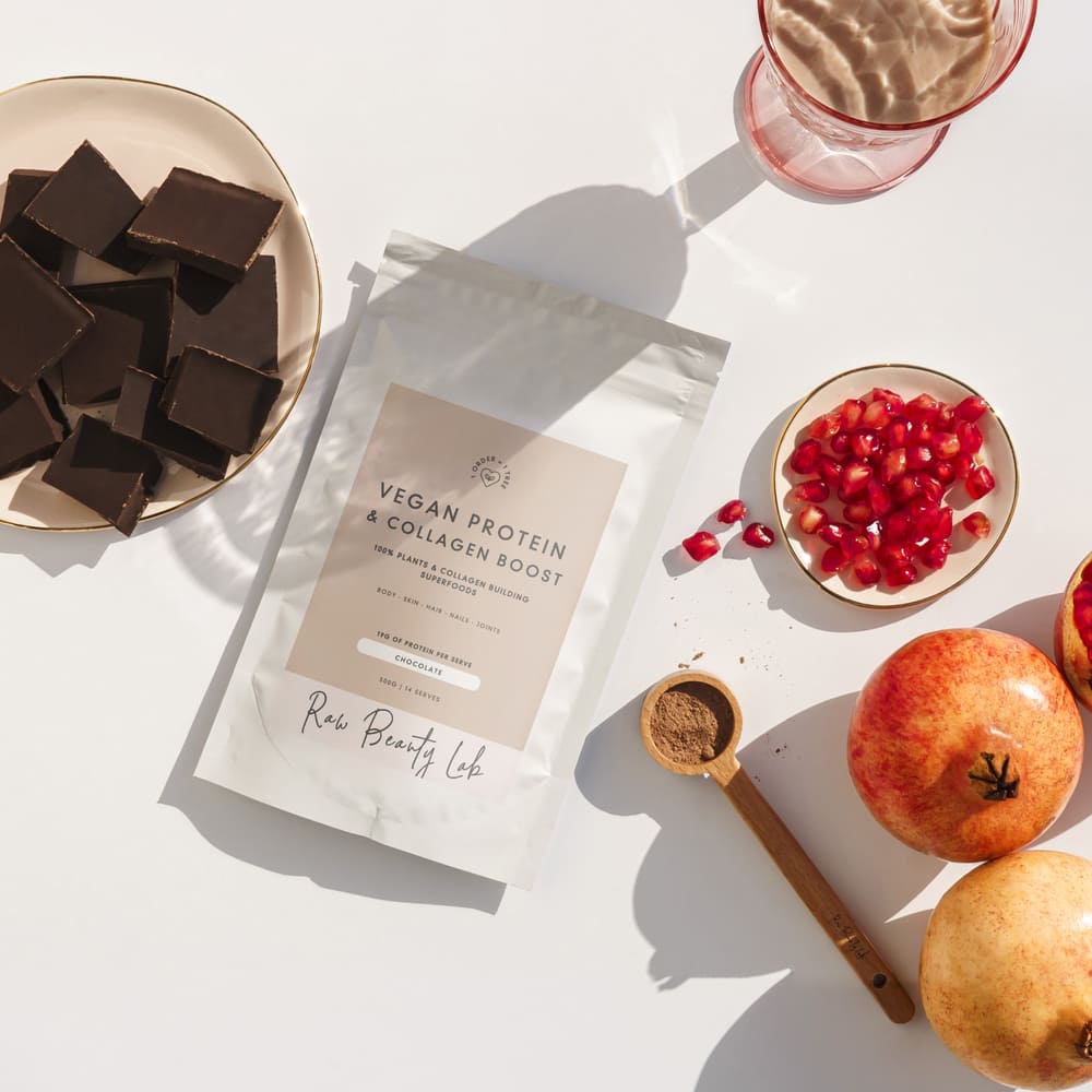 Flat lay of Raw Beauty Lab's Vegan Protein & Collagen Boost with chocolate squares, pomegranate seeds, pomegranates, a scoop of powder, and a glass of chocolate shake