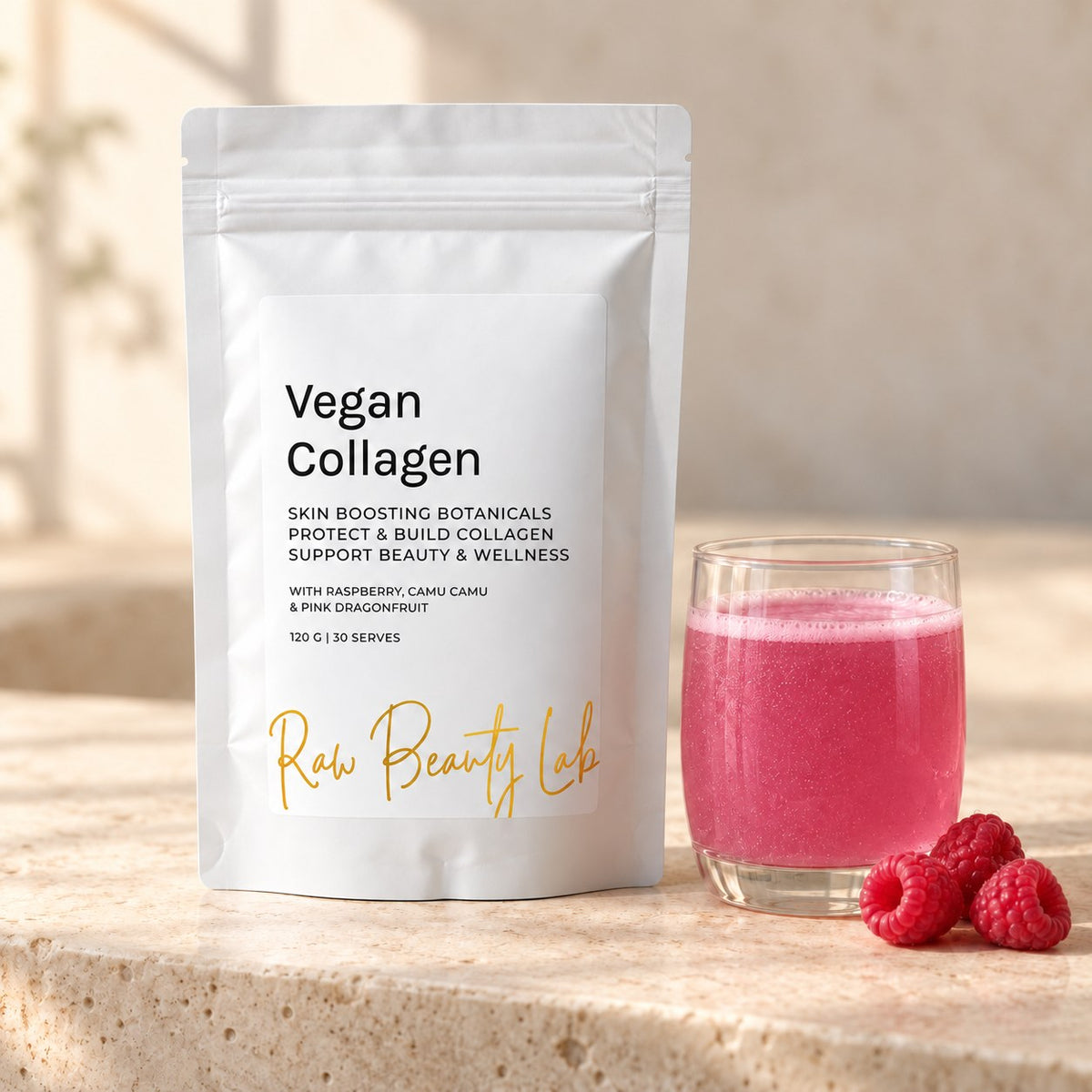 Vegan Collagen Booster pouch beside a glass of pink drink and raspberries on limestone